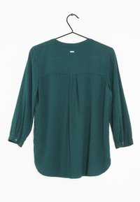 Teal långärmad skjorta i texturerad tyg, med en veckad bakdel, rund halsringning och knoppar vid ärmslut. Hänger från en svart krok.