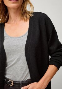 Zwarte gebreide cardigan met open voorkant, structuurstof en lange mouwen, gecombineerd met een grijs tanktop. Zwarte riem met een gouden gesp accent.