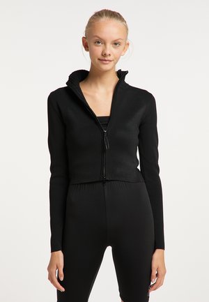 Ung kvinde med blondt hår iført en sort cropped trøje med lynlås og sorte højtaljede leggings mod en ensfarvet hvid baggrund.