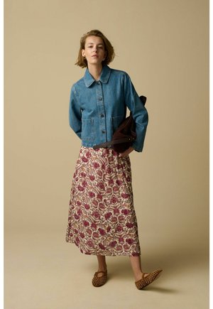 Jonge vrouw met een blauwe spijkerjas, een bloemenmidirok, bruine platgespijkerde schoenen en een donkerbruine tas tegen een beige achtergrond.