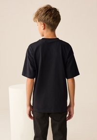 Tricou negru din bumbac, cu mâneci scurte, croială lejeră și decolteu rotund, prezentat din spate. Purtat cu blugi închiși la culoare.