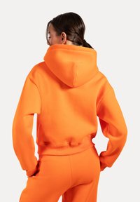 Haut à capuche orange en tissu doux avec une coupe décontractée. Doté d'une capuche avec cordon de serrage et de poignets côtelés, associé à un pantalon de jogger assorti.