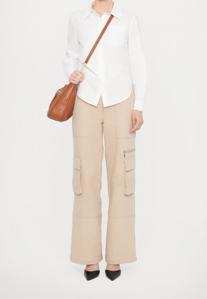 Donna che indossa una camicia bianca con bottoni e pantaloni cargo beige, con una borsa di pelle marrone con tracolla, e scarpe nere con punta appuntita.