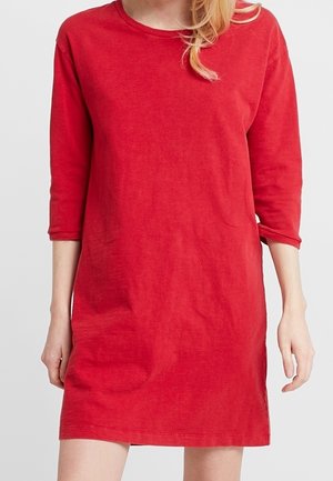 Kobieta ubrana w gładką, czerwoną sukienkę z długim rękawem, sięgającą do kolan, z okrągłym dekoltem, na neutralnym tle.