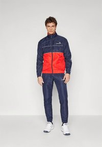 Jauns vīrietis stāv, ģērbies tumši zila un sarkana sporta jaku ar saskaņotām tumši zilām sporta biksēm un baltām sporta kurpēm, pret vienkāršu fonu.