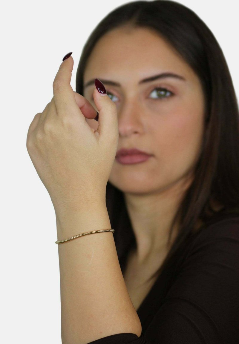 Bracciale d'oro, texture liscia, design flessibile, indossato attorno a un polso, mano sollevata con le dita puntate, smalto per unghie color bordeaux scuro, sfondo neutro.