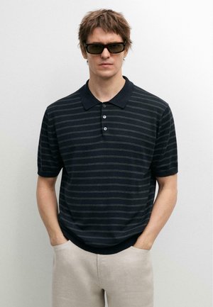 Mann trägt dunkel gestreiftes Polohemd, helle Hose und rechteckige Sonnenbrille, steht mit den Händen in den Taschen vor weißem Hintergrund.