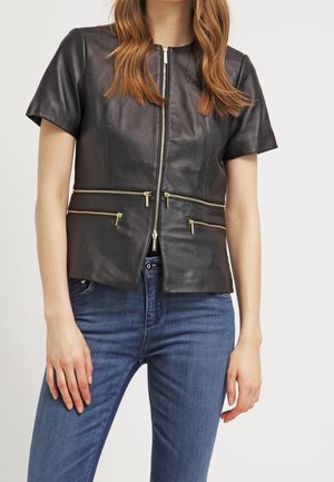 Frau trägt eine kurzärmelige schwarze Lederjacke mit mehreren goldenen Reißverschlüssen vorne, kombiniert mit blauen Denim-Jeans.