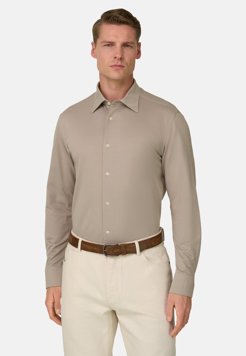 Homme portant une chemise beige à manches longues boutonnée, un pantalon crème et une ceinture en cuir marron, debout devant un fond clair uni.