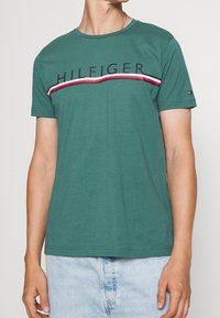 Camiseta de algodón verde con cuello redondo y mangas cortas, que presenta un logo de "HILFIGER" y franjas horizontales rojas, blancas y azules en el pecho.