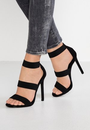 Füße, die schwarze Sandalen mit hohen Absätzen und drei Riemen tragen, kombiniert mit grauen Skinny Jeans, stehen auf einem hellen Boden.