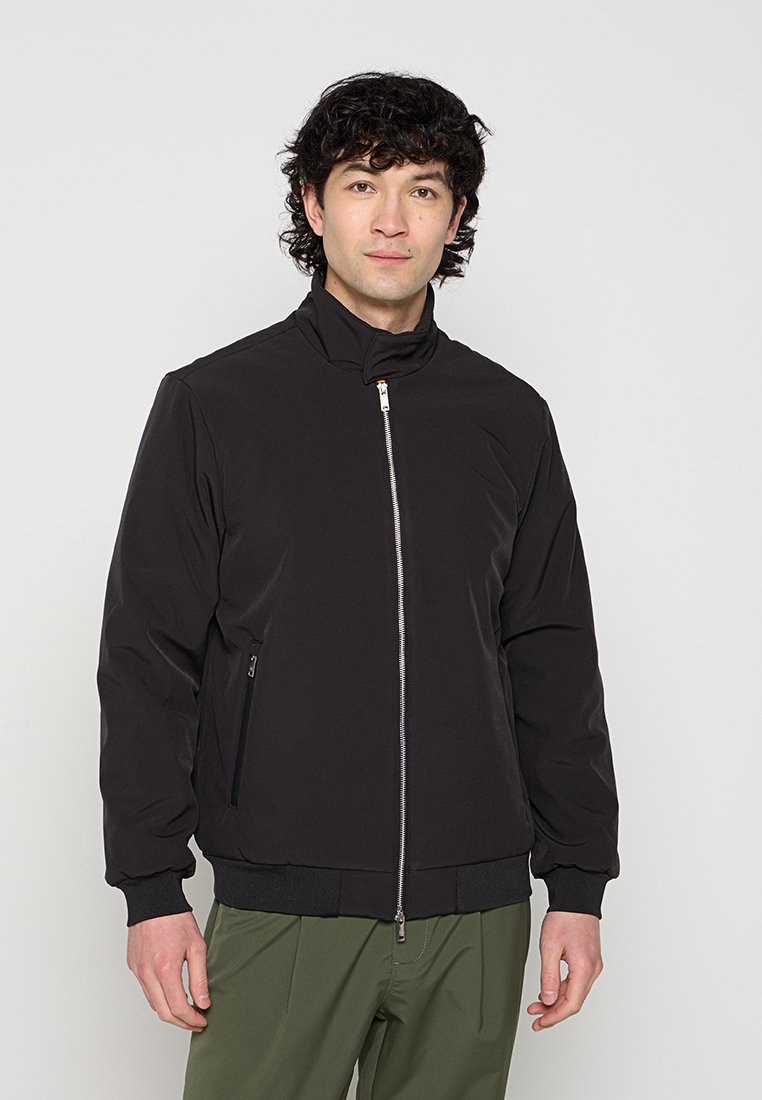 Jeune homme aux cheveux bouclés et foncés porte une veste noire zippée et un pantalon vert olive, debout devant un fond gris clair uni.