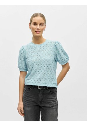 Kvinne iført lyseblå bluse med pufferm og hullmønster, og svarte jeans, stående foran en ensfarget hvit bakgrunn.