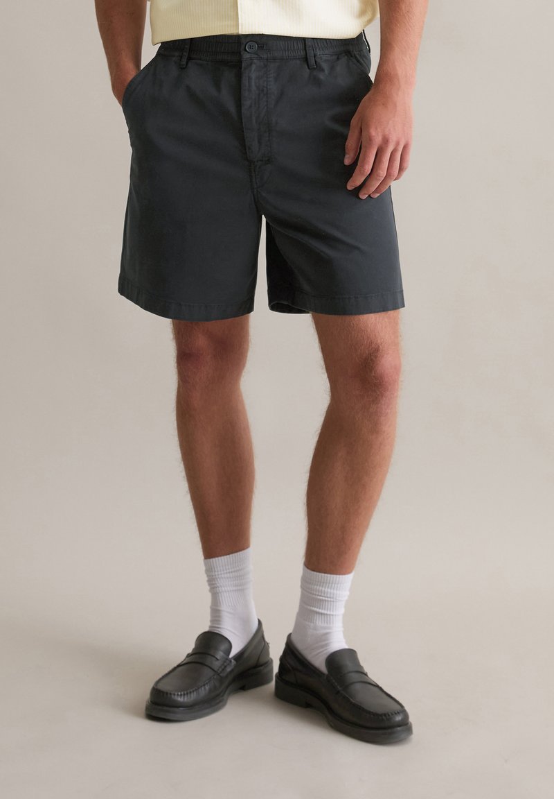 Zwarte katoenen shorts met zijzakken, een knoopsluiting en een relaxte pasvorm. Gdragen met witte sokken en zwarte loafers.