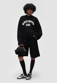 Czarny oversize sweter z białym napisem, czarne dopasowane szorty, czarne skarpety do kolan i srebrne trampki. Nosi małą czarną torebkę ze wzorem krokodyla.