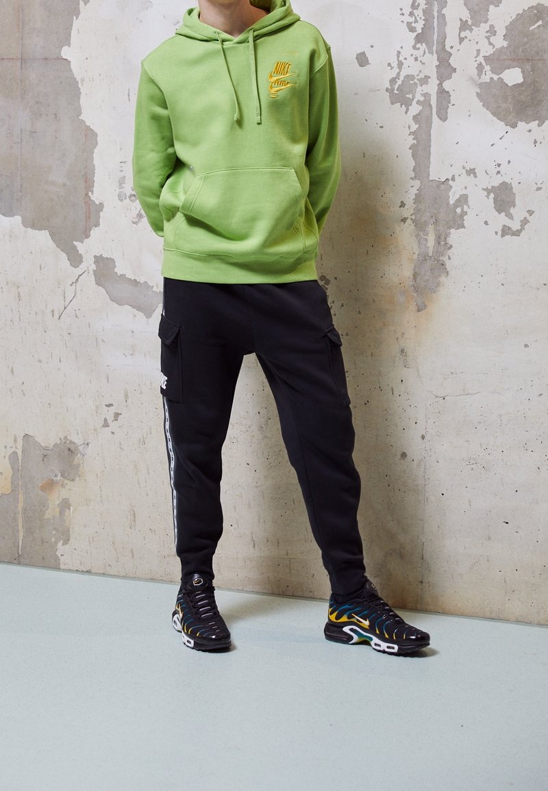 Grön zip hoodie med ficka framtill och gult logotyp, i kombination med svarta cargobyxor med sidofickor och vita detaljer. Svarta skor med en flerfärgad design.