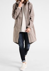 Giacca impermeabile beige chiaro con cappuccio, maniche lunghe e tasche con zip, abbinata a jeans scuri e sneaker chiare.