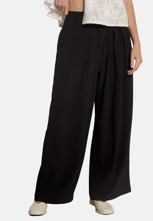 Kobieta nosząca czarne szerokie spodnie z wysokim stanem w połączeniu z białymi płaskimi butami i białym, teksturowanym, krótkim topem.