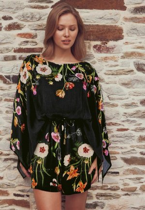 Woman wearing a black floral dress with wide sleeves standing in front of a textured white and brown brick wall with eyes closed.