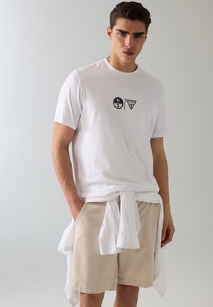 Giovane uomo che indossa una t-shirt bianca con stampa grafica, pantaloncini beige e una camicia bianca legata intorno alla vita, in piedi davanti a uno sfondo semplice.
