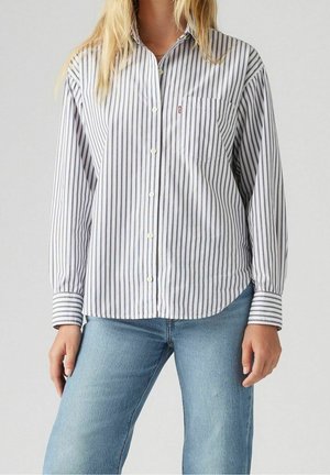 Femme portant une chemise blanche à manches longues avec de fines rayures verticales noires et un jean bleu clair, debout devant un fond uni.