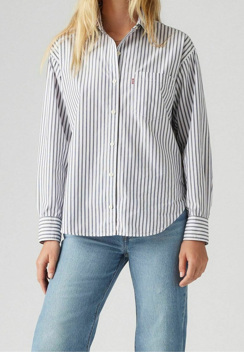 Femme portant une chemise blanche à manches longues avec de fines rayures verticales noires et un jean bleu clair, debout devant un fond uni.