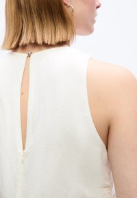 Πλάγια όψη ενός ατόμου με κοντά ξανθά μαλλιά που φοράει χρυσό κρίκο στο αυτί και λευκό αμάνικο τοπ με άνοιγμα τύπου κλειδαρότρυπα και κλείσιμο με φερμουάρ.