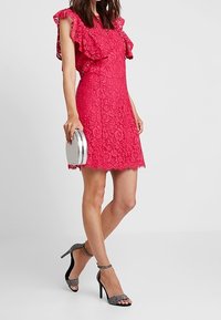 Rosa spetsklänning med volangärmar, scallopat fåll, ihopparad med svarta dekorerade högklackade skor och en silverfärgad rund handväska.