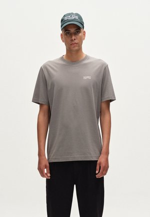 Gray cotton t-shirt with short sleeves, round neck, and small white logo. Paired with loose black pants and a dark green cap with white text.