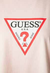 Šviesiai rožinė medvilninė marškinė su raudonu trikampiu logotipu, viduryje kurio užrašas "GUESS" ir klaustukas.