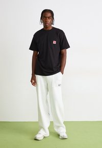 Black cotton T-shirt with a chest pocket featuring a red heart logo, paired with loose-fitting white sweatpants and grey sneakers.