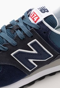 Modré a námořnické tenisky New Balance 574 se strukturovaným logem "N", modrými tkaničkami, síťovinou a bílou cedulkou s číslem 574 v červené barvě.