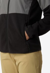 Fleeceová bunda se šedým kostkovaným horním dílem a černým dolním dílem. Obsahuje zipové kapsy a hladký, strukturovaný povrch.