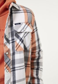 Primo piano di una persona che indossa una camicia a quadri con strisce arancioni, bianche e grigie sopra una felpa arancione.