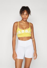 Soutien-gorge de sport jaune avec le texte blanc "NEBBIA", associé à un short blanc taille haute. Texture lisse, design minimaliste et coupe ajustée.