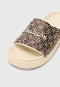 Brown patterned slide sandal with a textured strap, featuring "AIR JORDAN" branding in white. Soft cream outsole, open toe design.