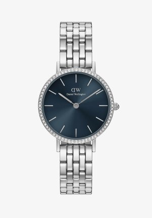 Hõbe käekell sinise kellapaneeli, kristallide aktsentidega äärise ja minimalistlike tunni märkidega. Brändi nimi on kuvatud 12 tunni positsiooni alla.