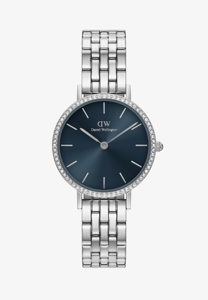 Hõbe käekell sinise kellapaneeli, kristallide aktsentidega äärise ja minimalistlike tunni märkidega. Brändi nimi on kuvatud 12 tunni positsiooni alla.