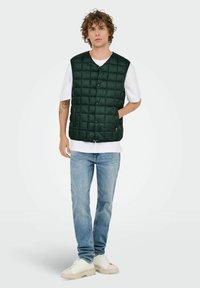 Gilet matelassé vert avec une fermeture à boutons, porté sur une chemise blanche, associé à un jean bleu et des baskets blanches ; texture lisse et design décontracté.