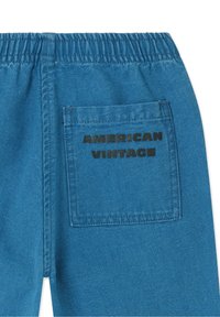 Pantalon en denim bleu avec une surface texturée et une taille élastique. Comprend une poche arrière avec l'inscription "AMERICAN VINTAGE" imprimée en noir.