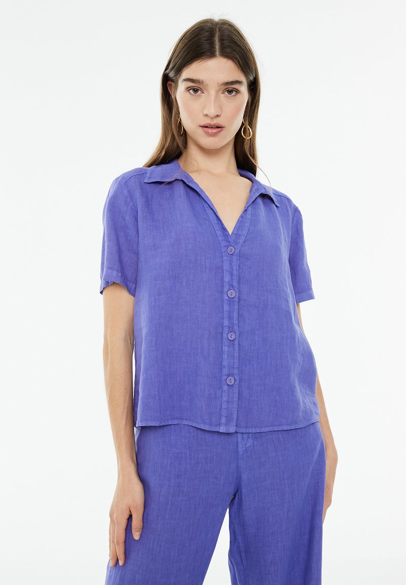 Camisa de botones de manga corta en lino morado vibrante, con cuello, escote en V y un ajuste relajado con un dobladillo ligeramente curvado.