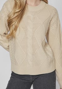 Beigefärgad stickad tröja med kabel- och diamantmönster, med en ribbad halsringning och fåll. Bärs tillsammans med mörkgrå jeans.