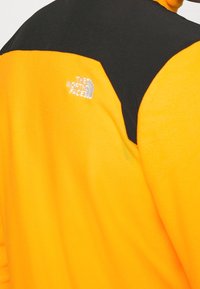 Giacca in pile con corpo arancione e area spalla nera. Presenta un logo riflettente argentato sul lato sinistro superiore. Texture morbida.