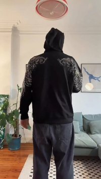 Osoba v černé mikině s kapucí s bílými spirálovými vzory na rukávech a zádech stojí v obývacím pokoji a dívá se od kamery.