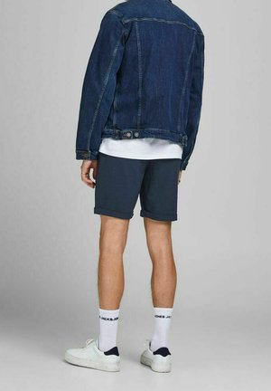 Giacca di jeans blu scuro con maniche lunghe, t-shirt bianca sotto, pantaloncini blu navy, scarpe sportive bianche e calzini crew con marchio.