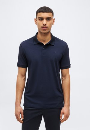 Junger Mann mit kurzen Haaren, der ein eng anliegendes dunkelblaues Poloshirt und dunkle Hosen trägt, vor einem einfarbigen hellen Hintergrund stehend.