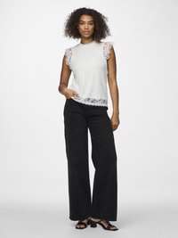 Femeie cu păr creț, purtând un top alb fără mâneci din dantelă, blugi negri cu picioare largi și sandale negre cu toc, stând cu o mână în buzunar.