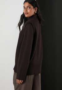 Dark brown knitted sweater with a high neck, ribbed cuffs, and textured finish, paired with light brown pinstripe trousers.