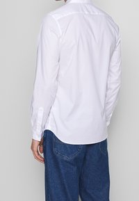 Camisa blanca de manga larga con cuello abotonado, canesú trasero y dobladillo ligeramente ancho, combinada con vaqueros de mezclilla, vista desde atrás.