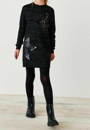 Mujer con un vestido de suéter negro de manga larga con dos grandes estrellas de lentejuelas, medias negras y botines negros caminando en interiores.